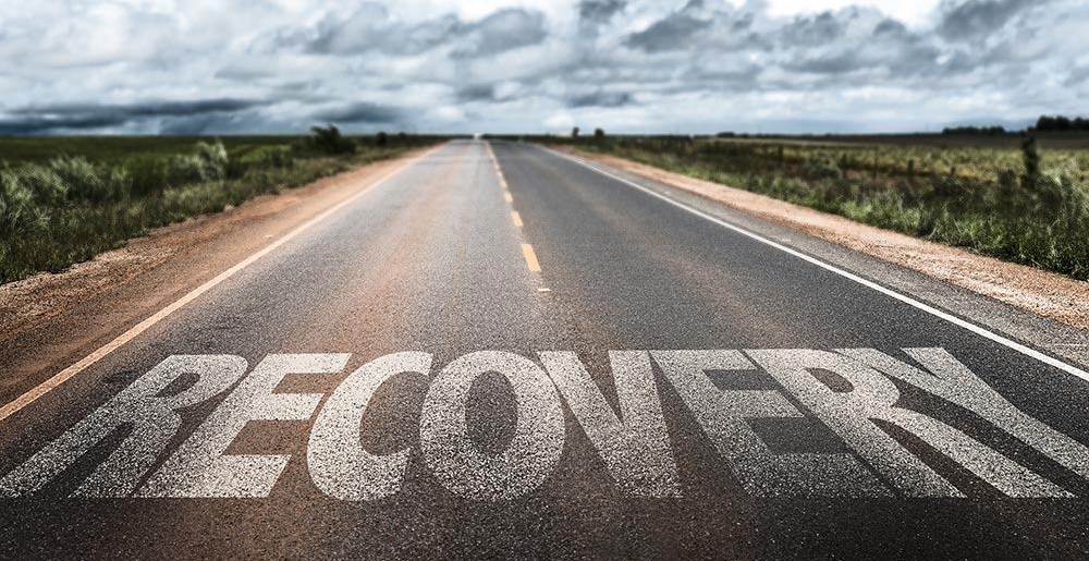 An image of a road with the word 'recovery' painted on it, symbolizing the journey from incarceration to the community amid the challenges explored in the blog.