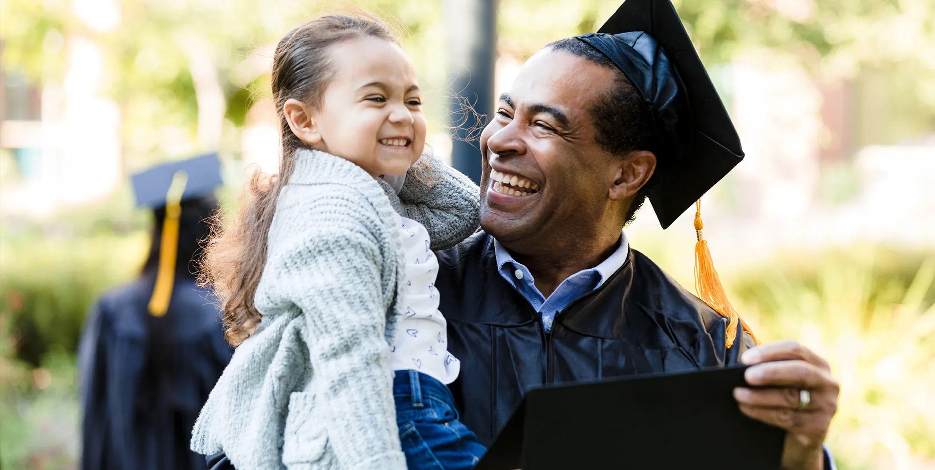 A formerly incarcerated man with his daughter in his arms dons the cap and gown, proudly receiving his high school equivalency diploma.