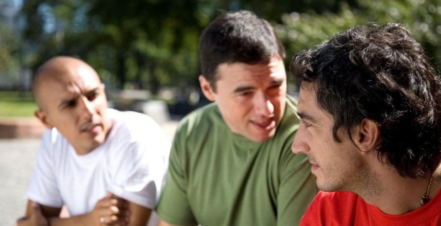Three young men in street clothes talk closely on a city sidewalk, showing peer pressure in a group.